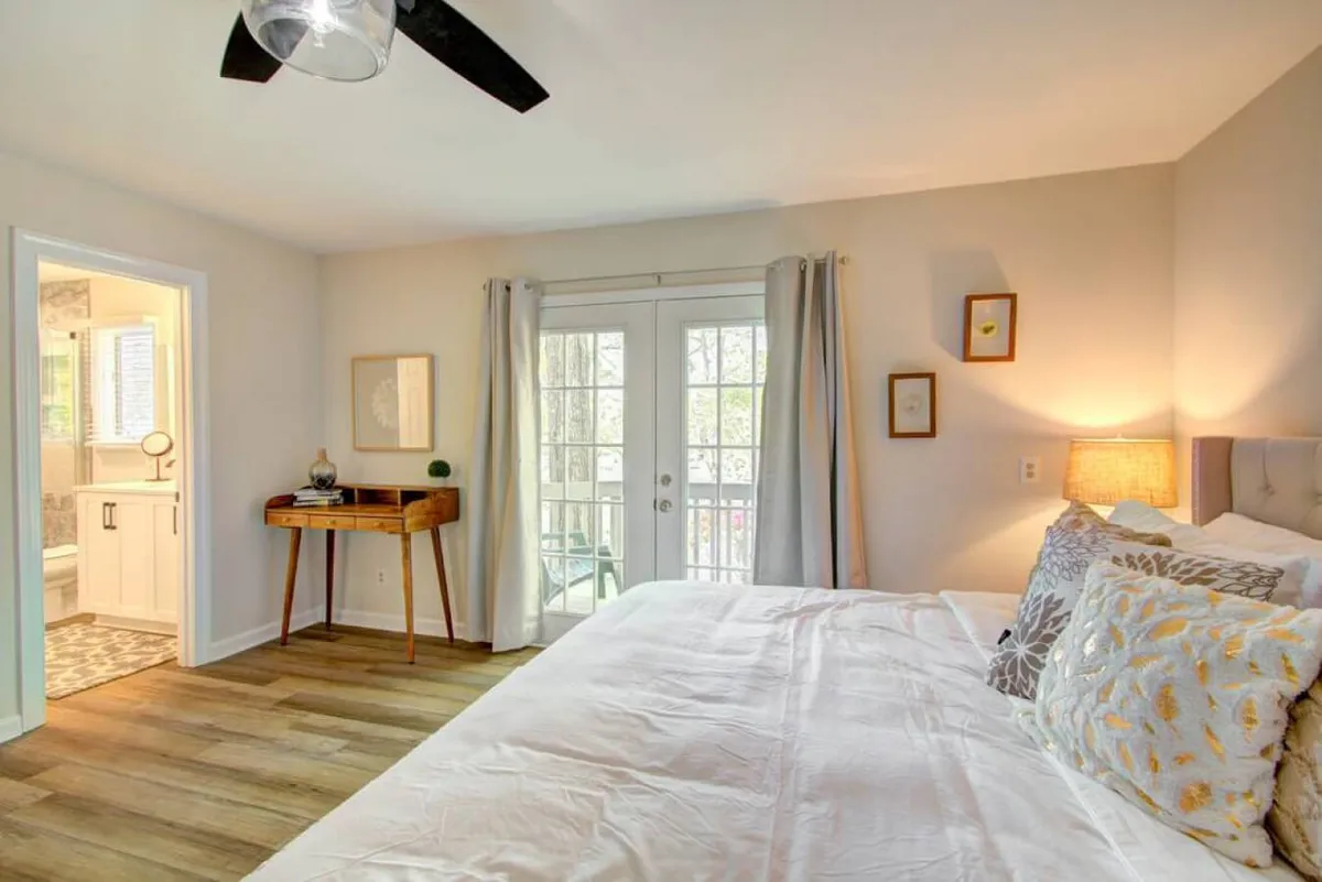 A bright bedroom with a comfortable bed adorned with decorative pillows, French doors that open to a private balcony, and a wooden side table. The room is filled with natural light, offering a calm yet inviting environment ideal for restful nights and peaceful mornings.