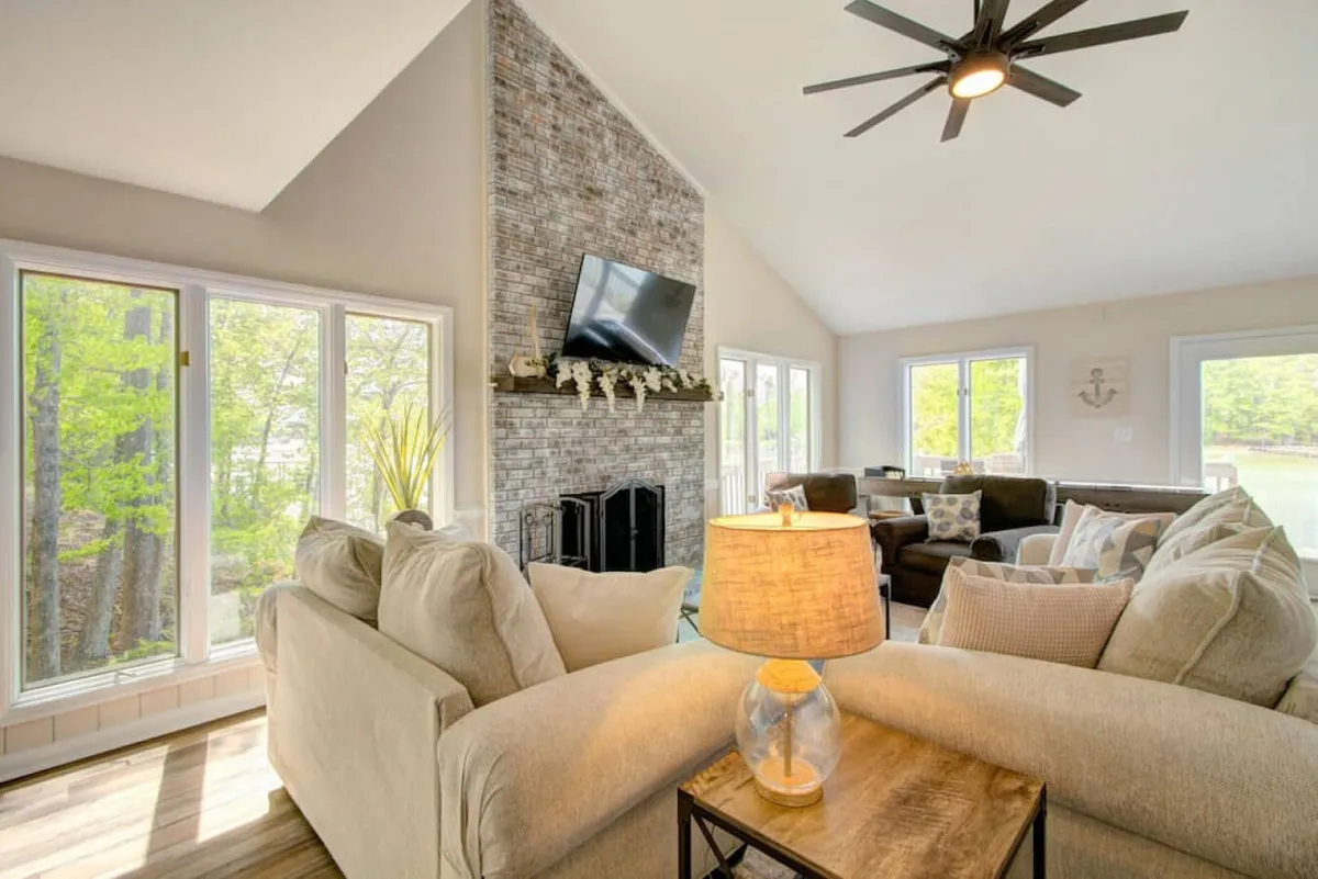 A spacious living room featuring a large seating area, a stone fireplace, and a TV mounted on the wall. Multiple large windows allow abundant sunlight and provide uninterrupted views of the lake, enhancing the cozy and welcoming mood of the room.
