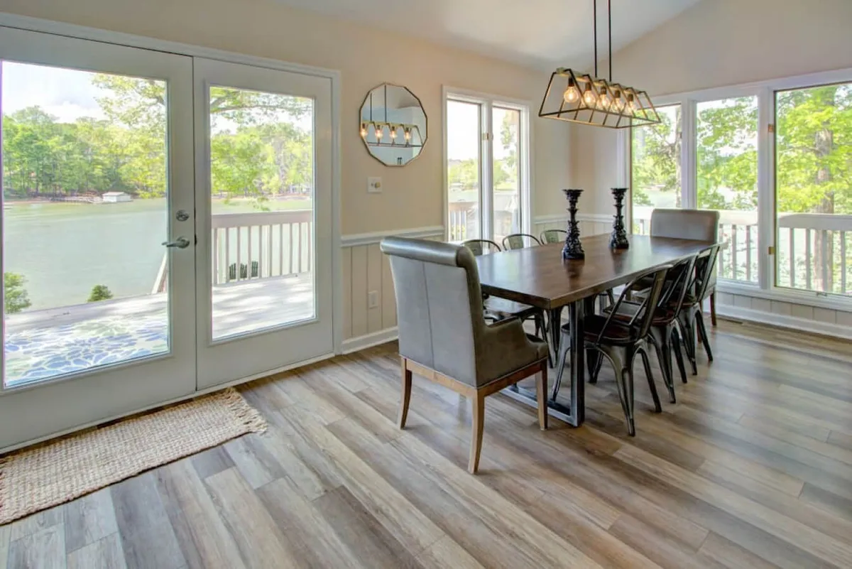 A warmly lit dining area with a large dining table set beneath an elegant chandelier. Expansive windows frame stunning outdoor water views, and natural light pours in, illuminating the heart of the open space. The room feels inviting, perfect for memorable meals and group gatherings.