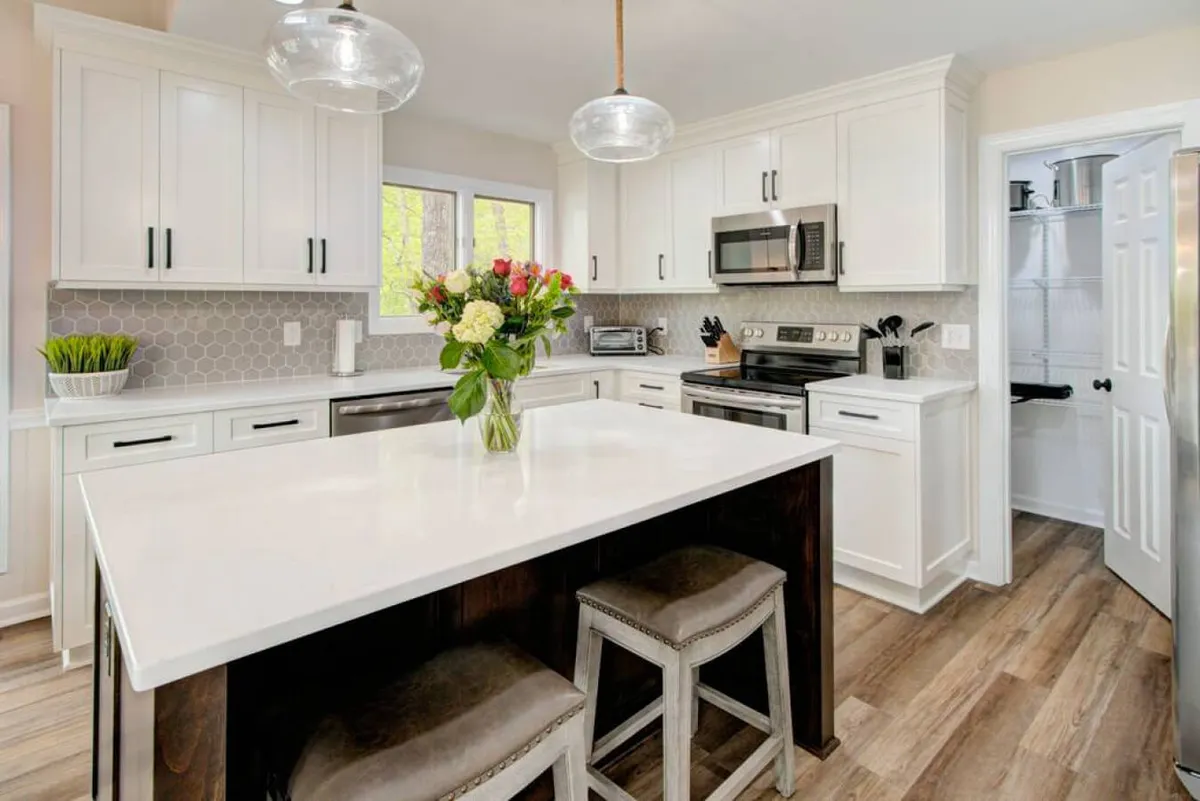 A spacious kitchen featuring a white island with ample seating, a flower vase for a touch of freshness, and sleek stainless steel appliances lining the back wall. The room is filled with natural light, highlighting the generous workspace and inviting setup ideal for social cooking and gatherings.