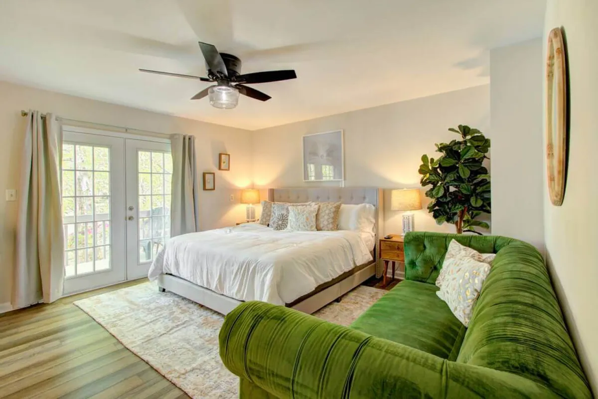 A spacious, sunlit main bedroom featuring a king-sized bed with decorative pillows, a plush green velvet sofa, and large windows that bathe the room in natural light. An indoor plant adds a touch of freshness, and the overall ambiance is tranquil and inviting.