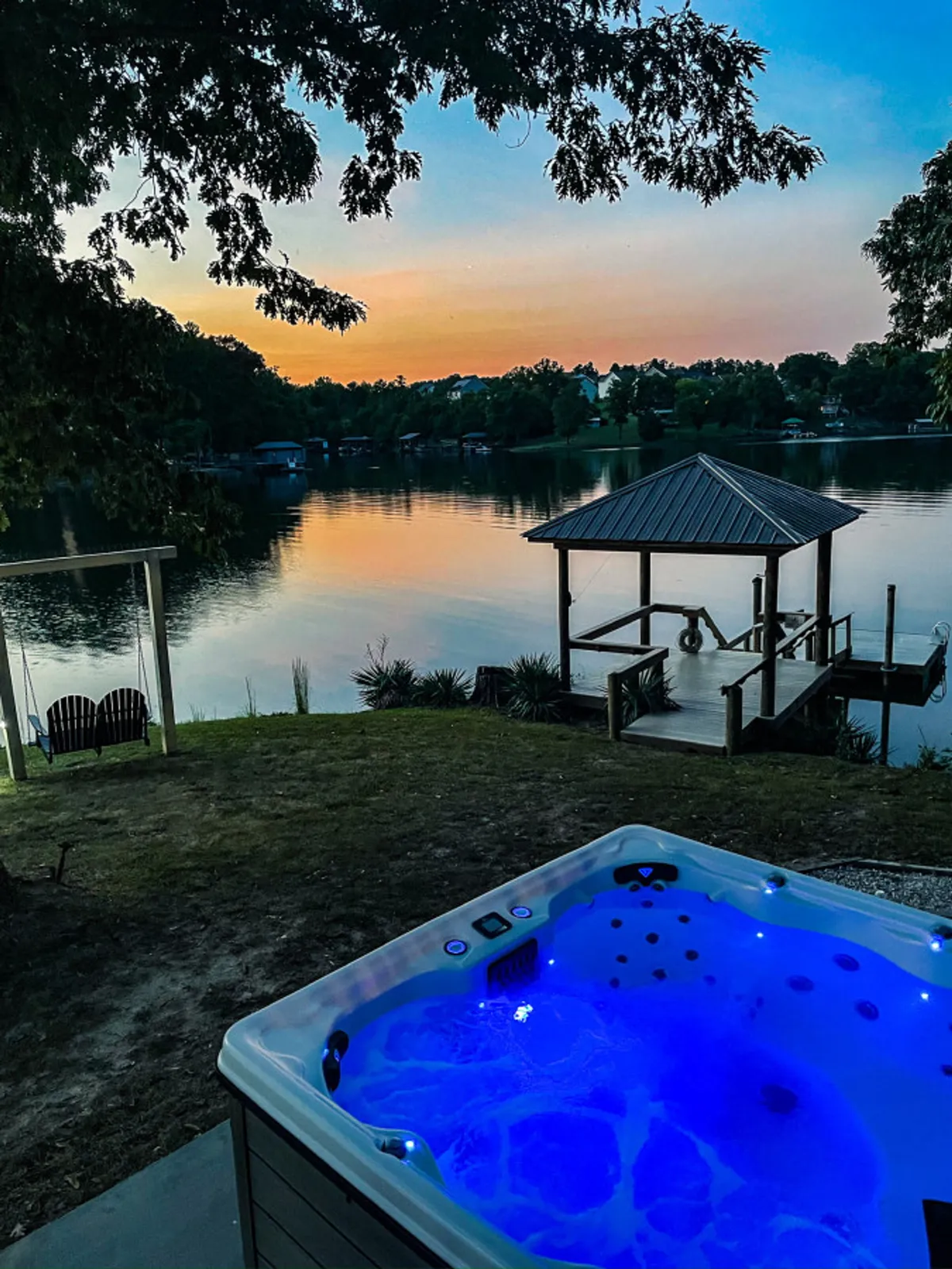 A panoramic shot featuring a spacious hot tub in the foreground, a private dock leading out onto a calm lake, and the sky ablaze with sunset hues. The image includes the dock and lake, suggesting access to watercraft and outdoor adventure, while the hot tub area is ready for guests to enjoy.