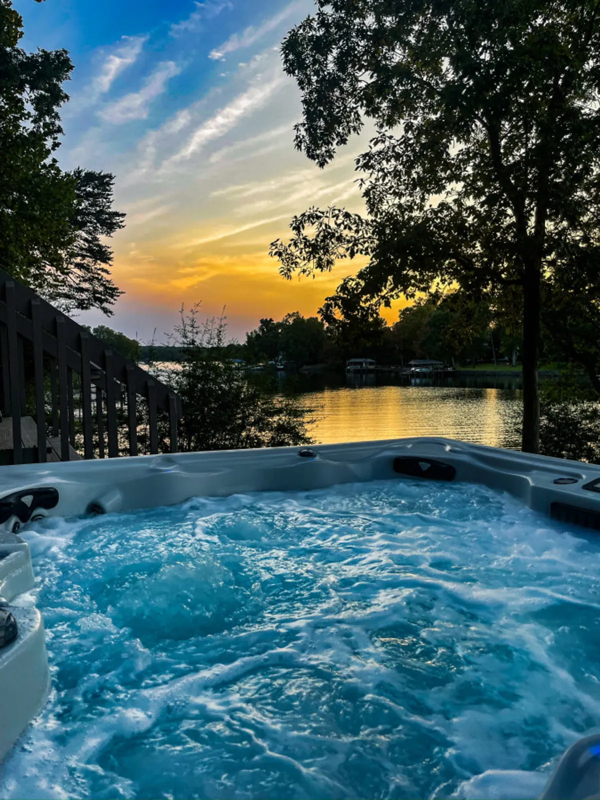 An inviting, steamy 8-person hot tub sits on a deck overlooking a tranquil lake, framed by sunset colors and leafy trees. Friends are relaxing in the tub, enjoying the warmth and the stunning lake view. The atmosphere is serene and social, with the comfort of the hot tub highlighted against the backdrop of the natural surroundings.