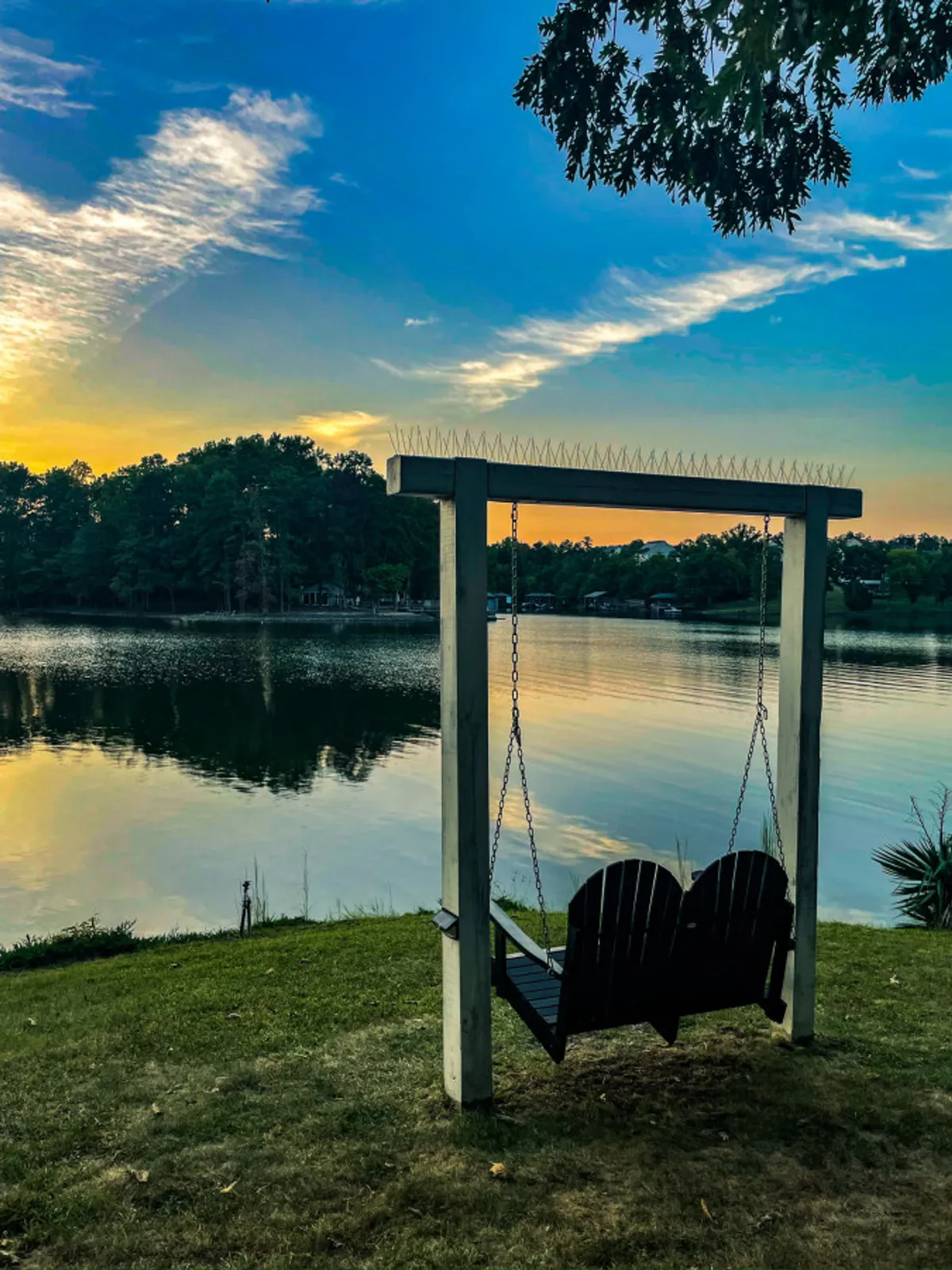 An inviting swing faces out over the lake, framed by lush trees and a glowing sunset. The tranquil surroundings encourage quiet contemplation, relaxation, and shared memories while embracing the peacefulness of the cove setting.